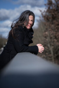 Frau mit schwarzen Haaren und in schwarzem Mantel lehnt an einem Geländer. Es ist Herbst. Dunkle Wolken und Wind in den Haaren